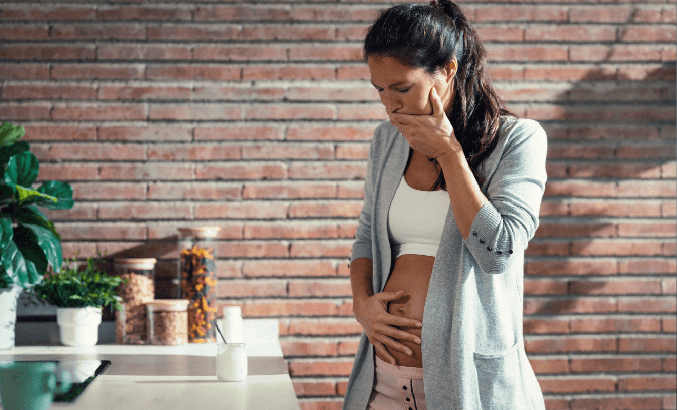 Pregnant woman experiencing nausea while standing in a kitchen, holding her stomach and covering her mouth.