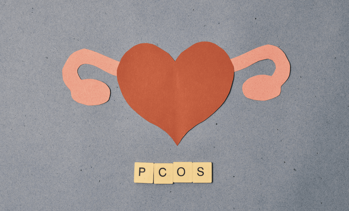 Paper cutout illustration of female reproductive organs shaped like a heart, symbolizing PCOS and fertility awareness, with wooden tiles spelling ‘PCOS’ on a textured background