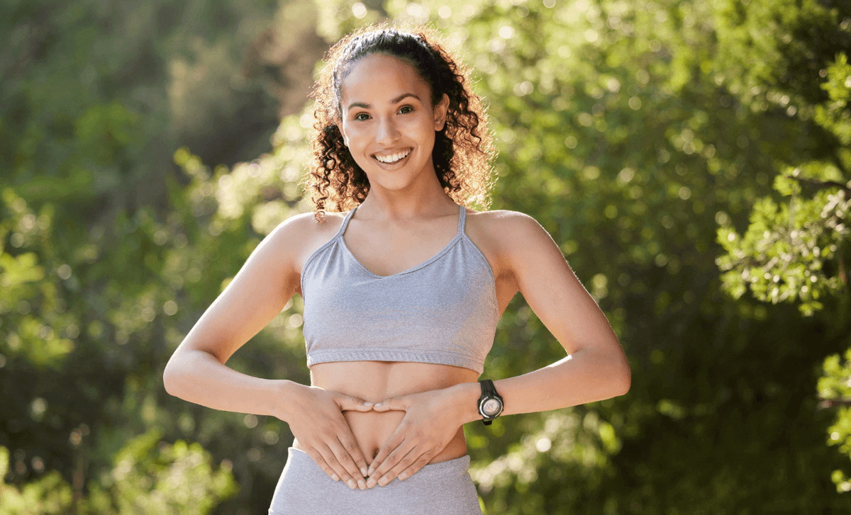 Smiling woman in athletic wear outdoors, gently holding her belly to symbolize natural pregnancy preparation, healthy lifestyle, and fertility wellness.