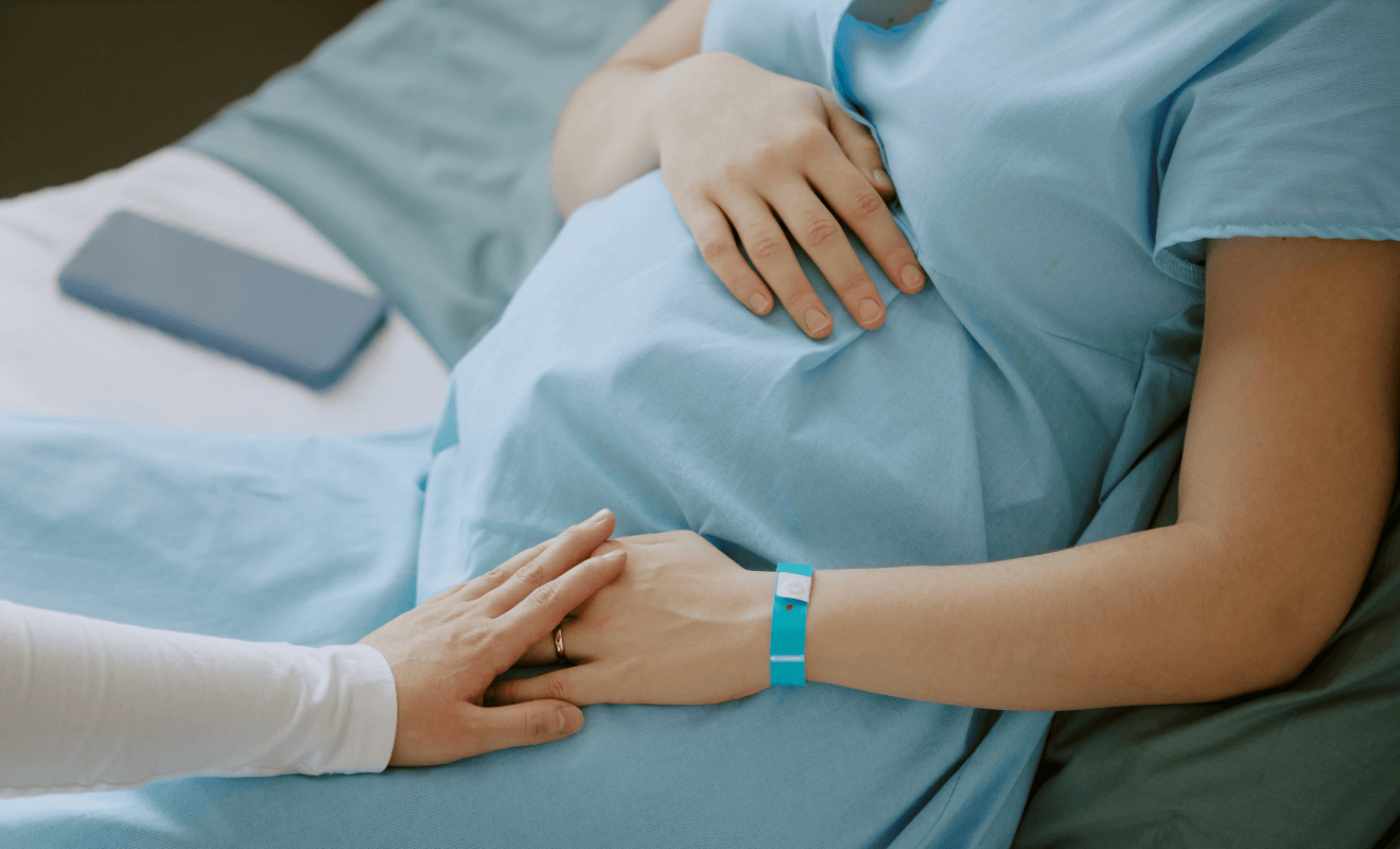 A pregnant woman in a hospital gown lying on a bed, gently cradling her baby bump while holding hands with a supportive partner or healthcare provider. 