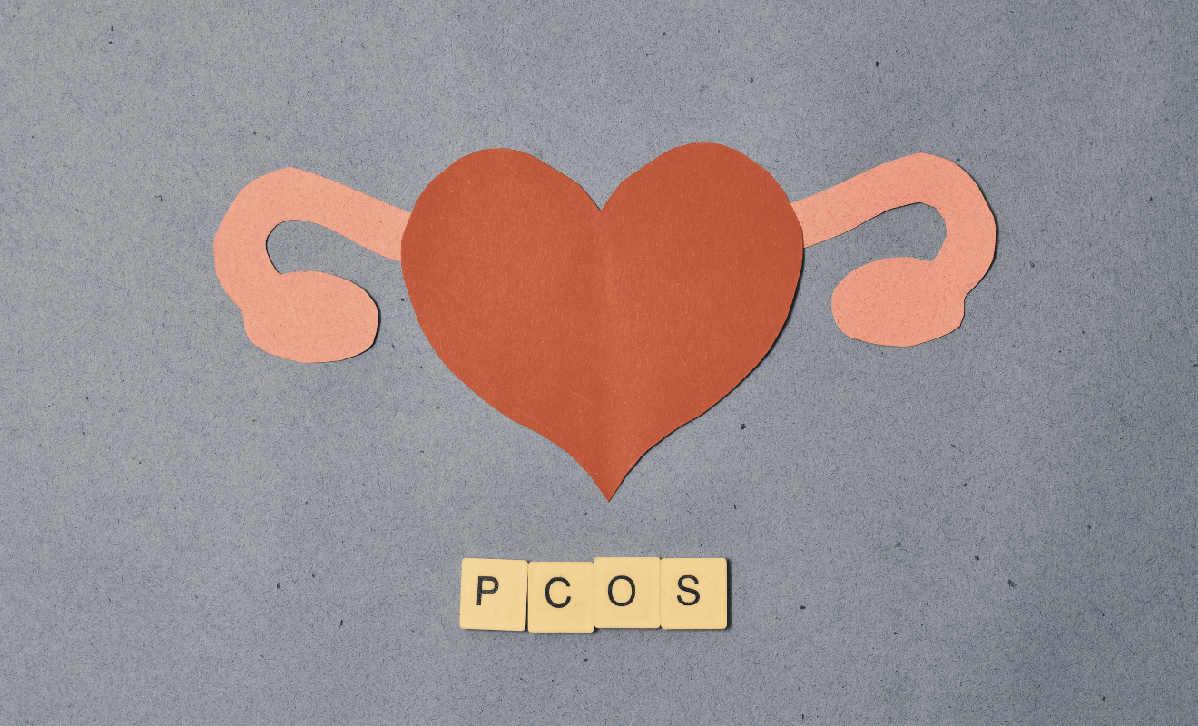 Paper cutout illustration of female reproductive organs shaped like a heart, symbolizing PCOS and fertility awareness, with wooden tiles spelling ‘PCOS’ on a textured background