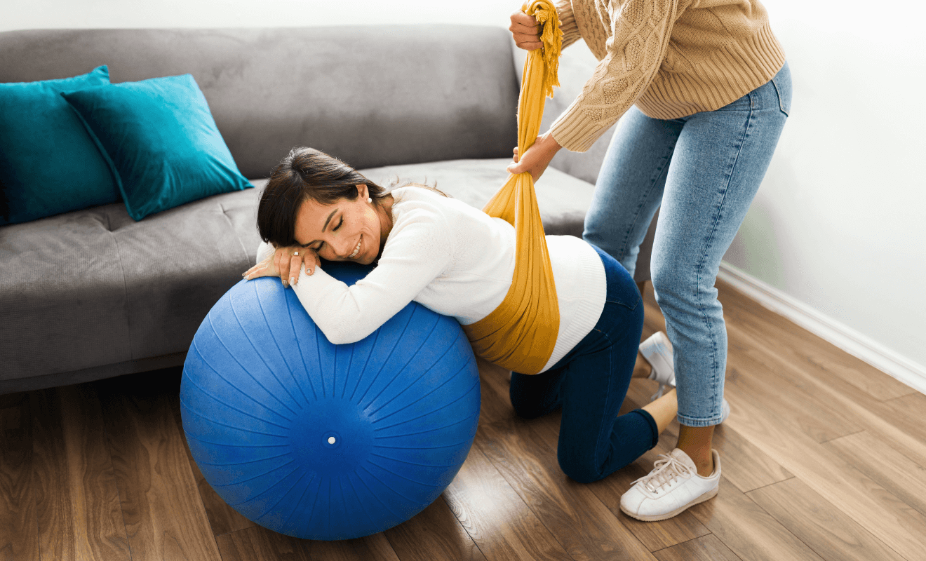 Pregnant woman using a birthing ball at home with support from a doula using a rebozo technique for pain relief during labor.