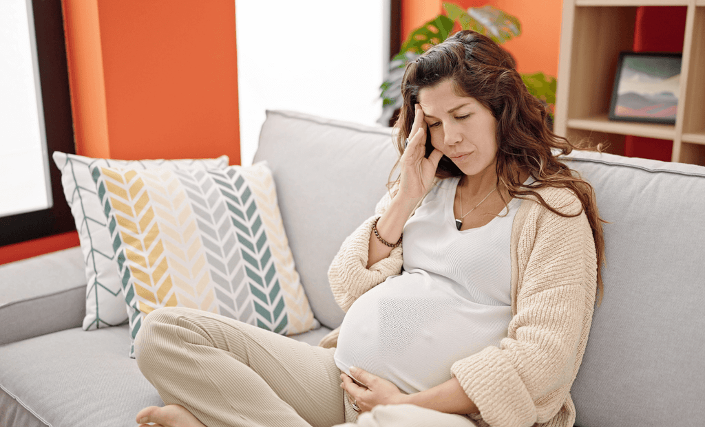 Pregnant woman sitting on a couch, looking concerned while touching her forehead, indicating stress or discomfort.
