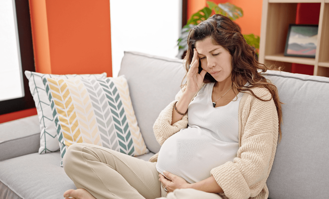 Pregnant woman sitting on a couch, looking concerned while touching her forehead, indicating stress or discomfort.