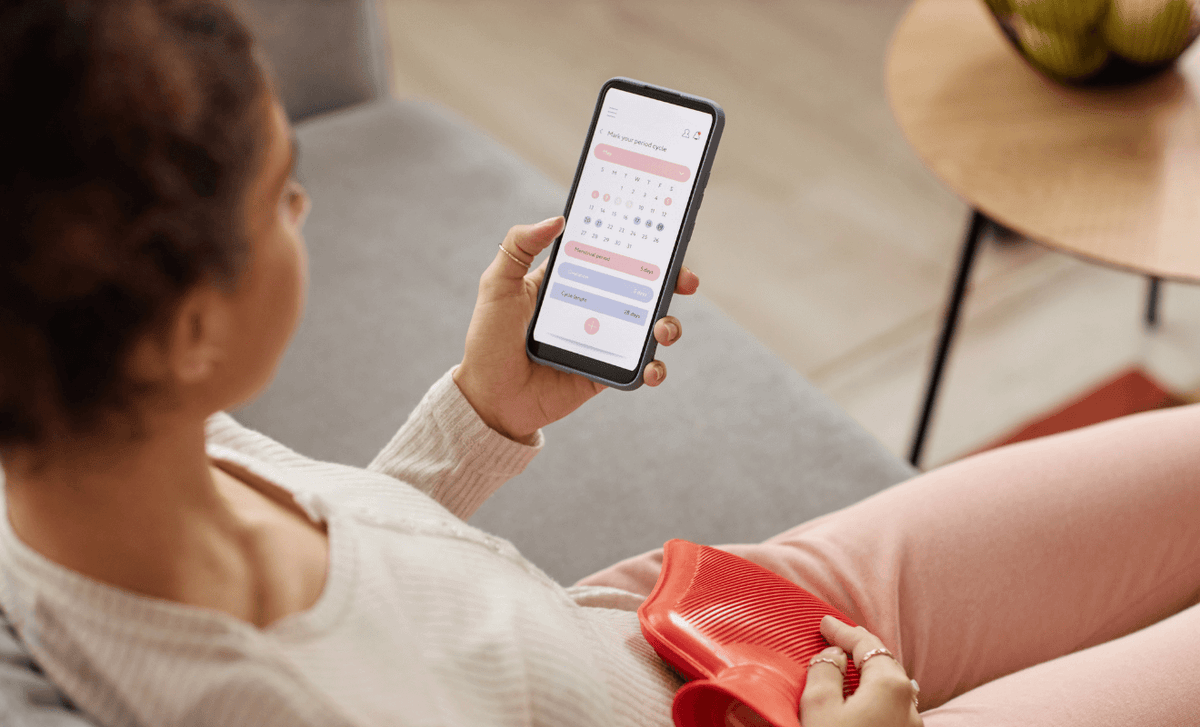 Woman on a sofa checking a cycle tracking app on her phone, with a red hot water bottle in her lap