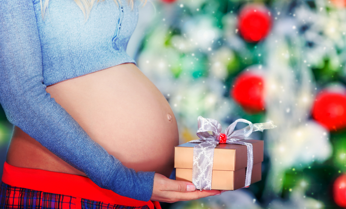 Pregnant woman holding a wrapped holiday gift in front of a Christmas tree, symbolizing thoughtful holiday gifts for expecting moms.