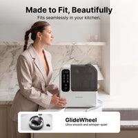 Woman beside the Momcozy D8 bottle washer on a kitchen counter, showing its wheeled base and drain hose