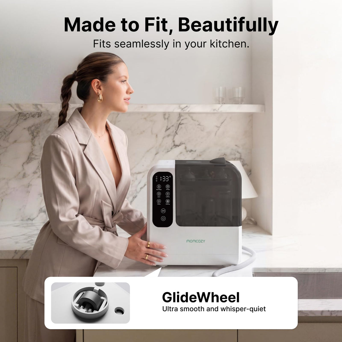 Woman beside the Momcozy D8 bottle washer on a kitchen counter, showing its wheeled base and drain hose