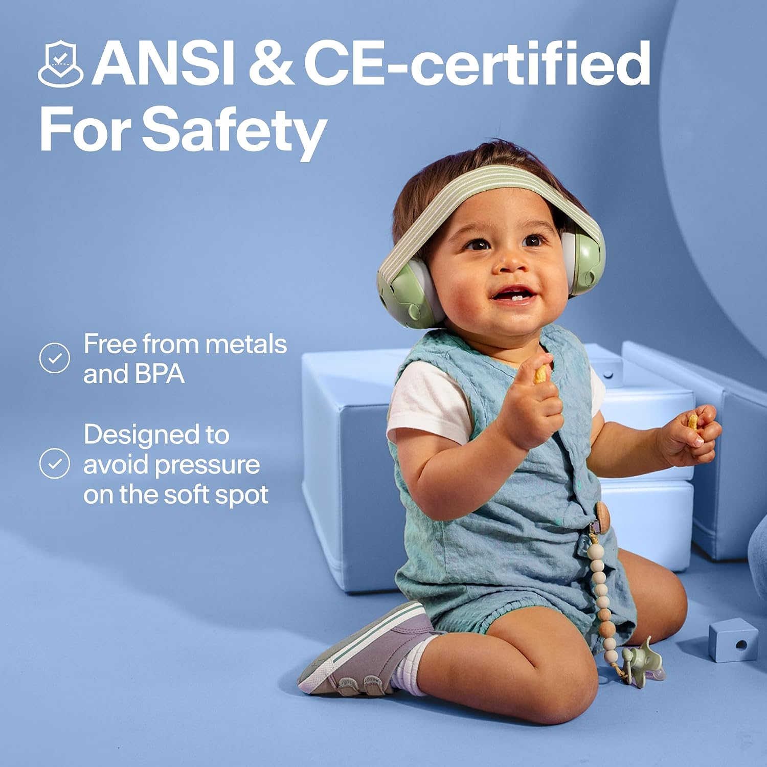 Baby sitting up wearing sage green Alpine Muffy Baby earmuffs, with notes that they are free from metals and BPA and designed to avoid pressure on the soft spot