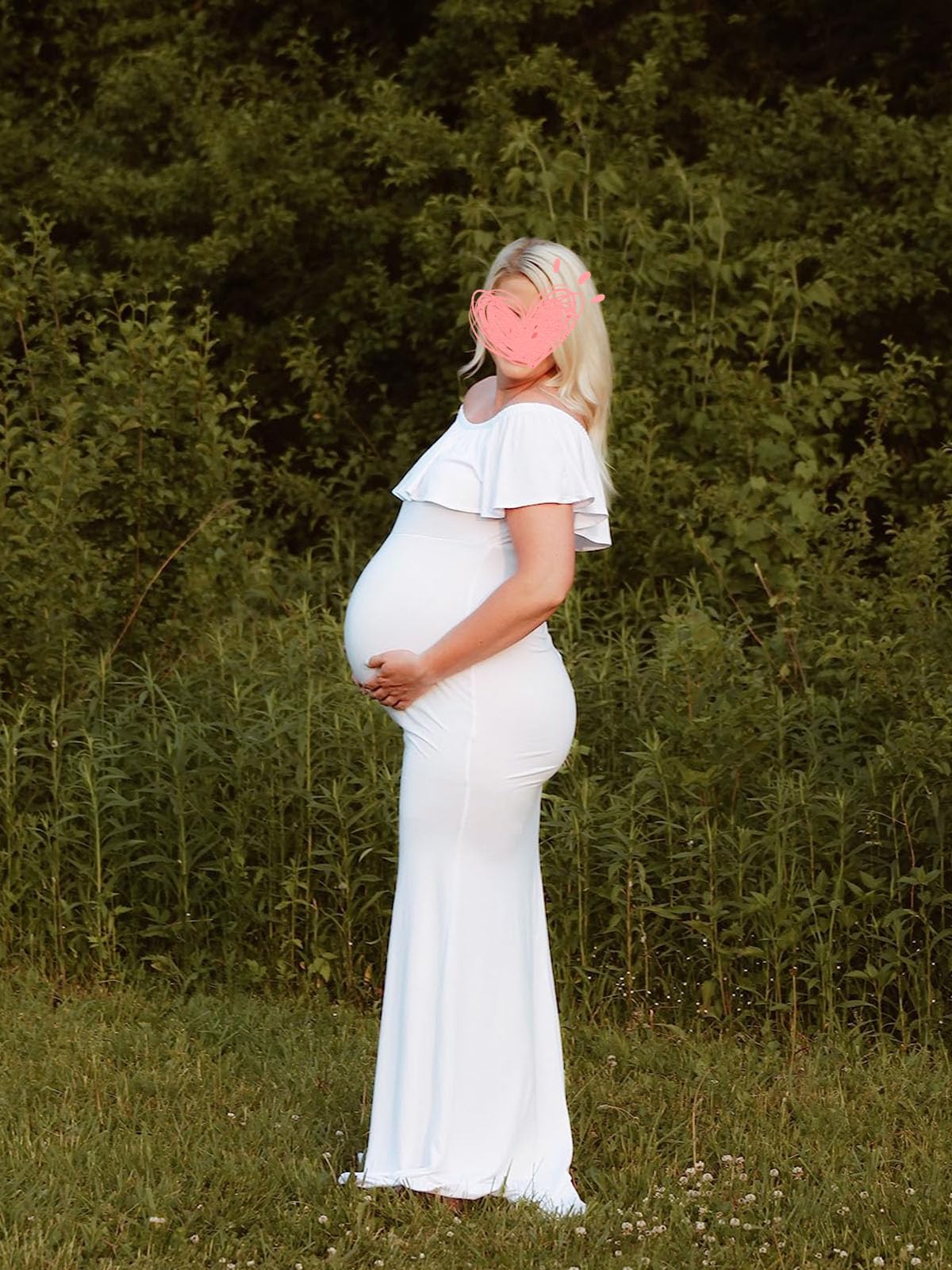 Maternity photoshoot dress in white worn by a pregnant woman standing outdoors with greenery in the background.