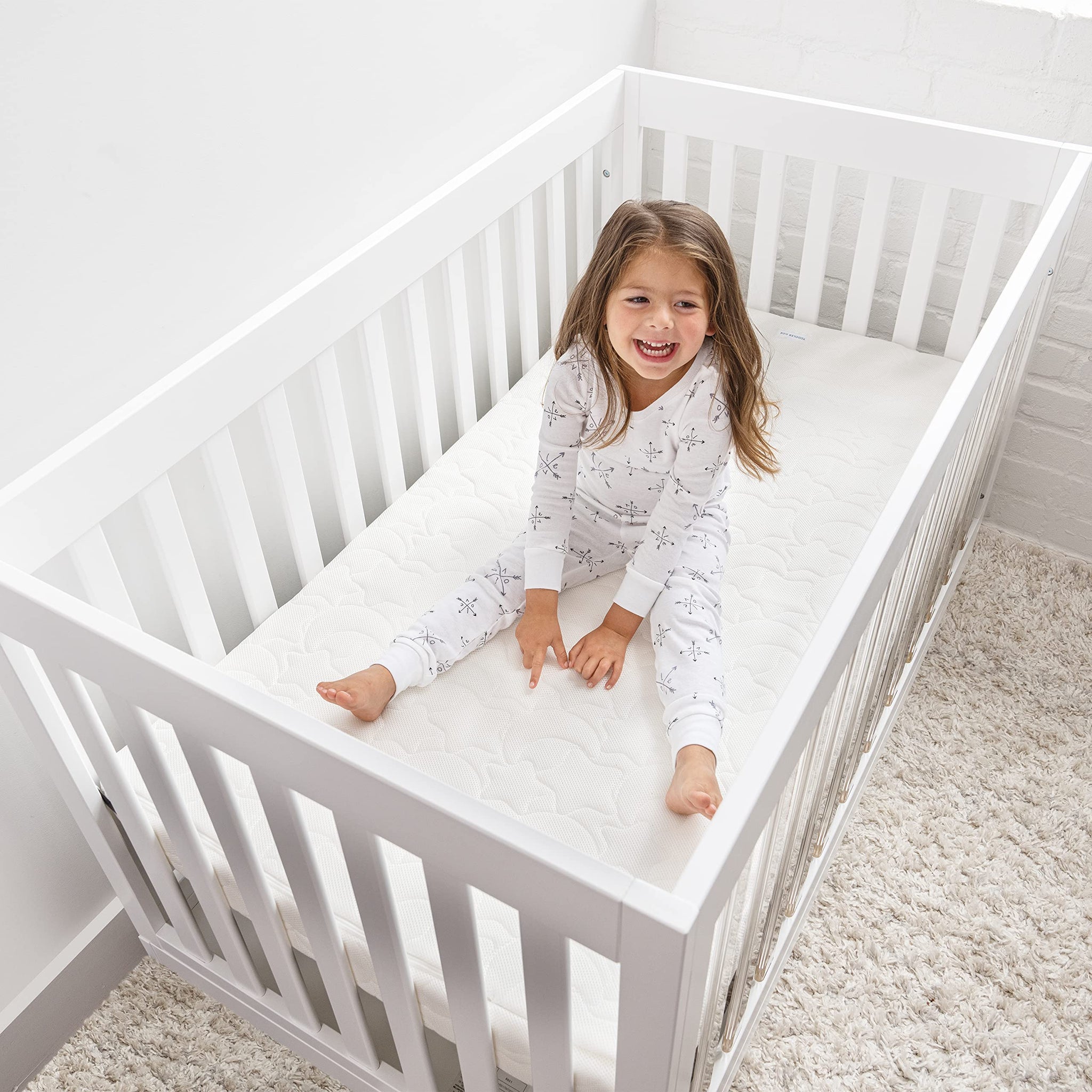 Happy child sitting on HALO DreamWeave breathable crib mattress in a white crib, showcasing comfort and safety for sleep.