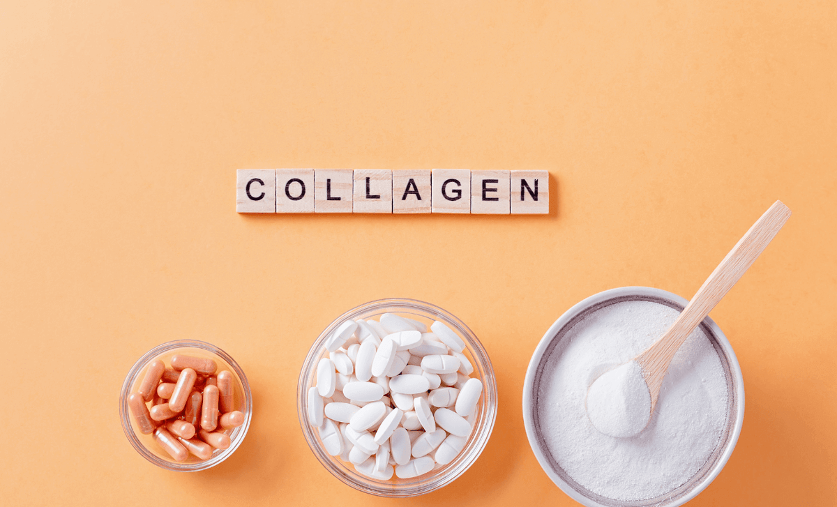 Letter tiles spelling COLLAGEN above glass bowls of collagen capsules, tablets and powder with a wooden scoop on an orange background